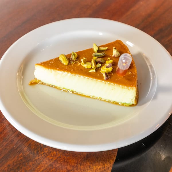 Slice of flan with caramel sauce, pistachios, and a honey dropper on a white plate.